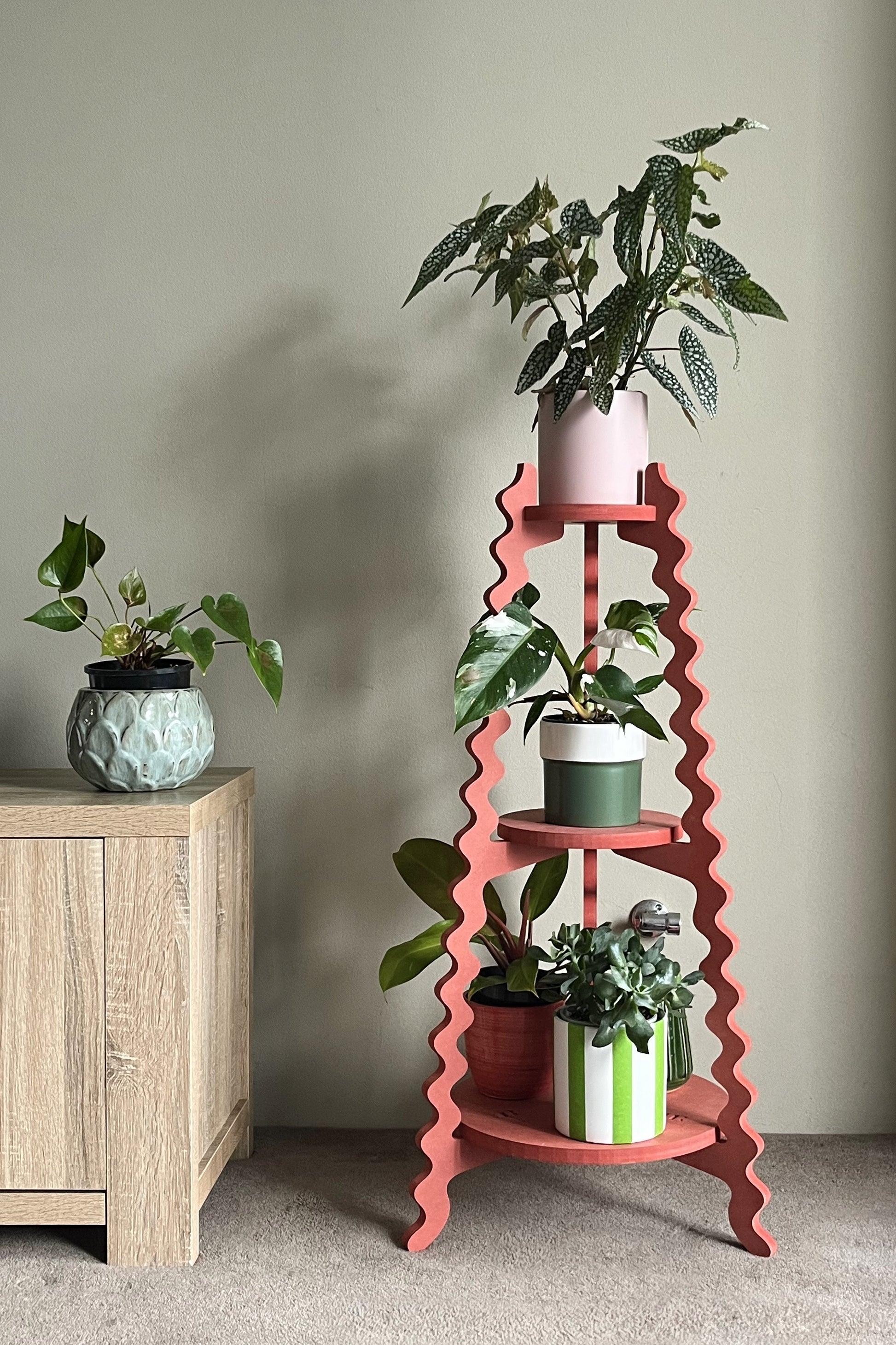A red wavy Xigglie Poppy Threezy Plant Stand  supporting potted plants on three tiers against a neutral wall.