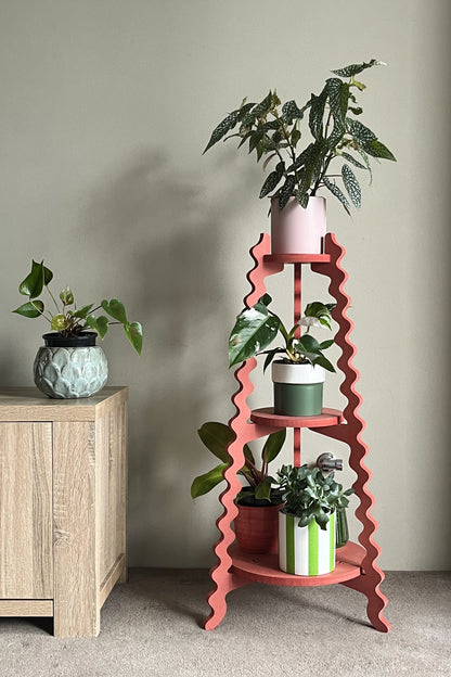 A red wavy Xigglie Poppy Threezy Plant Stand  supporting potted plants on three tiers against a neutral wall.
