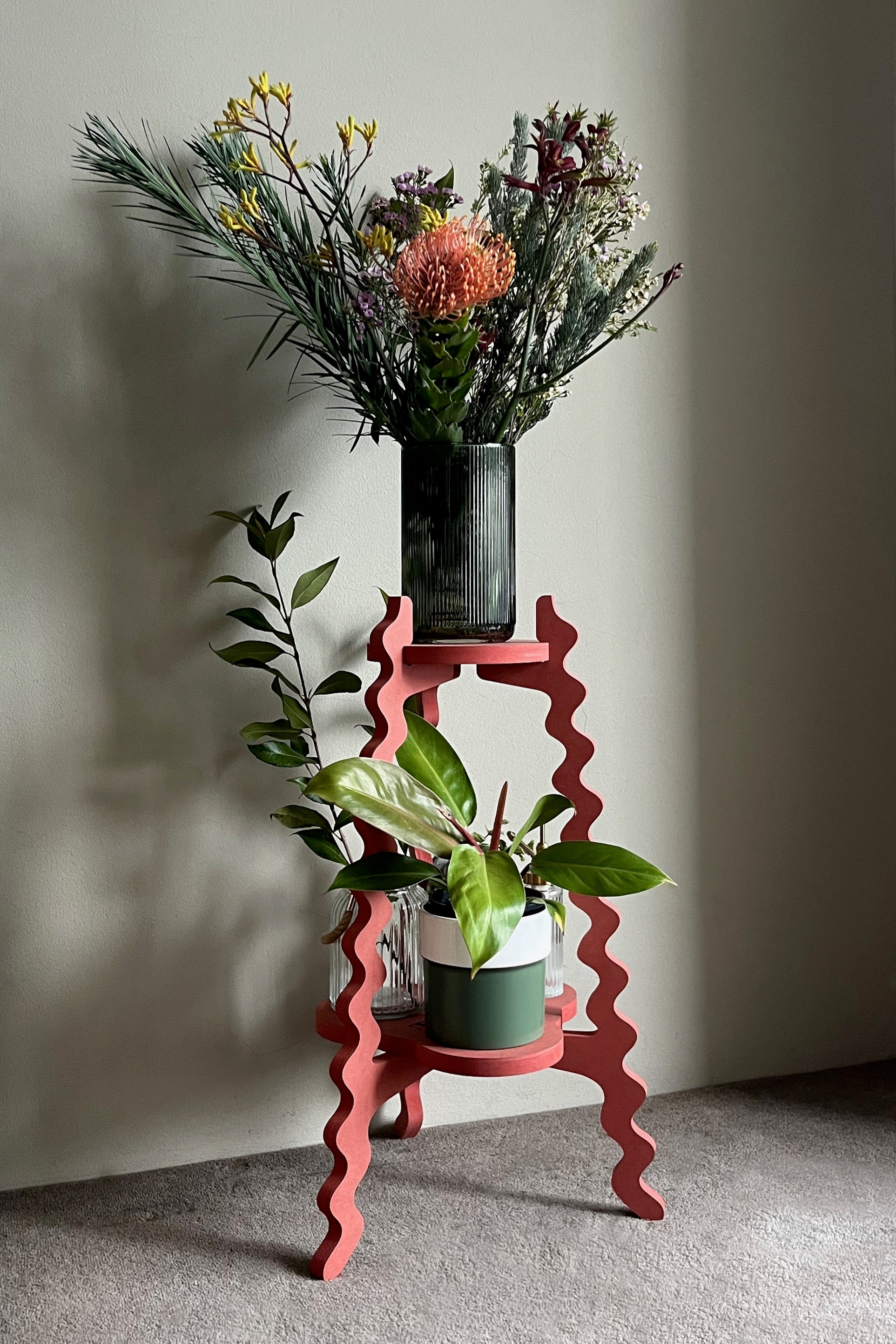 Red wavy Xigglie Poppy Two-UP Plant Stand with a vase of flowers and potted plants against a plain wall.