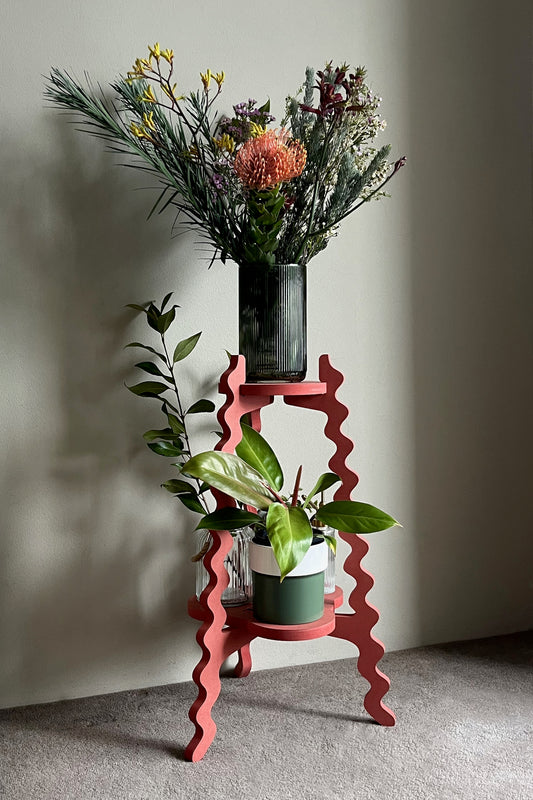 Red wavy Xigglie Poppy Two-UP Plant Stand with a vase of flowers and potted plants against a plain wall.