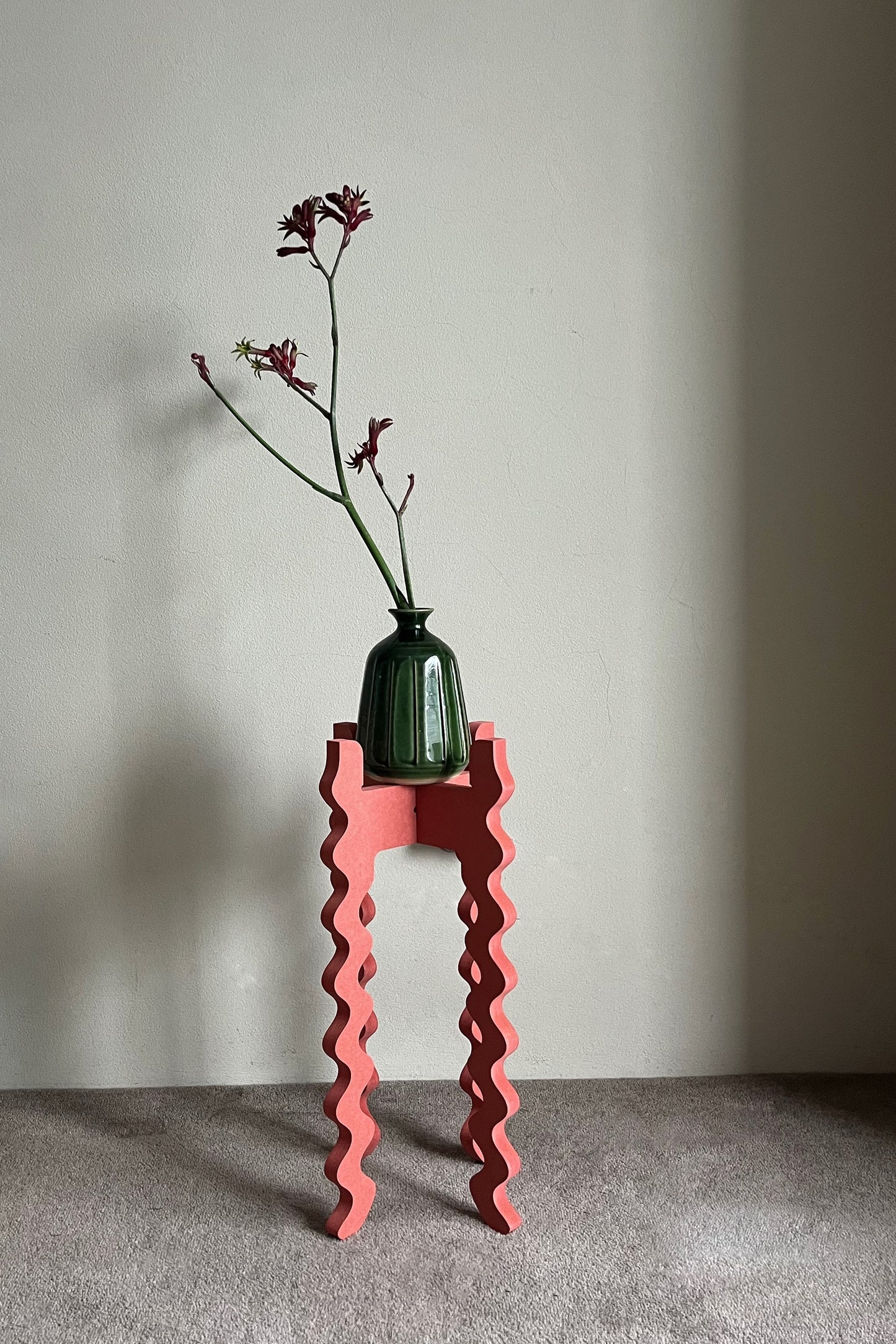 Green vase with a plant on a red wavy Xigglie Poppy Tallboy Wide Plant Stand against a plain wall.