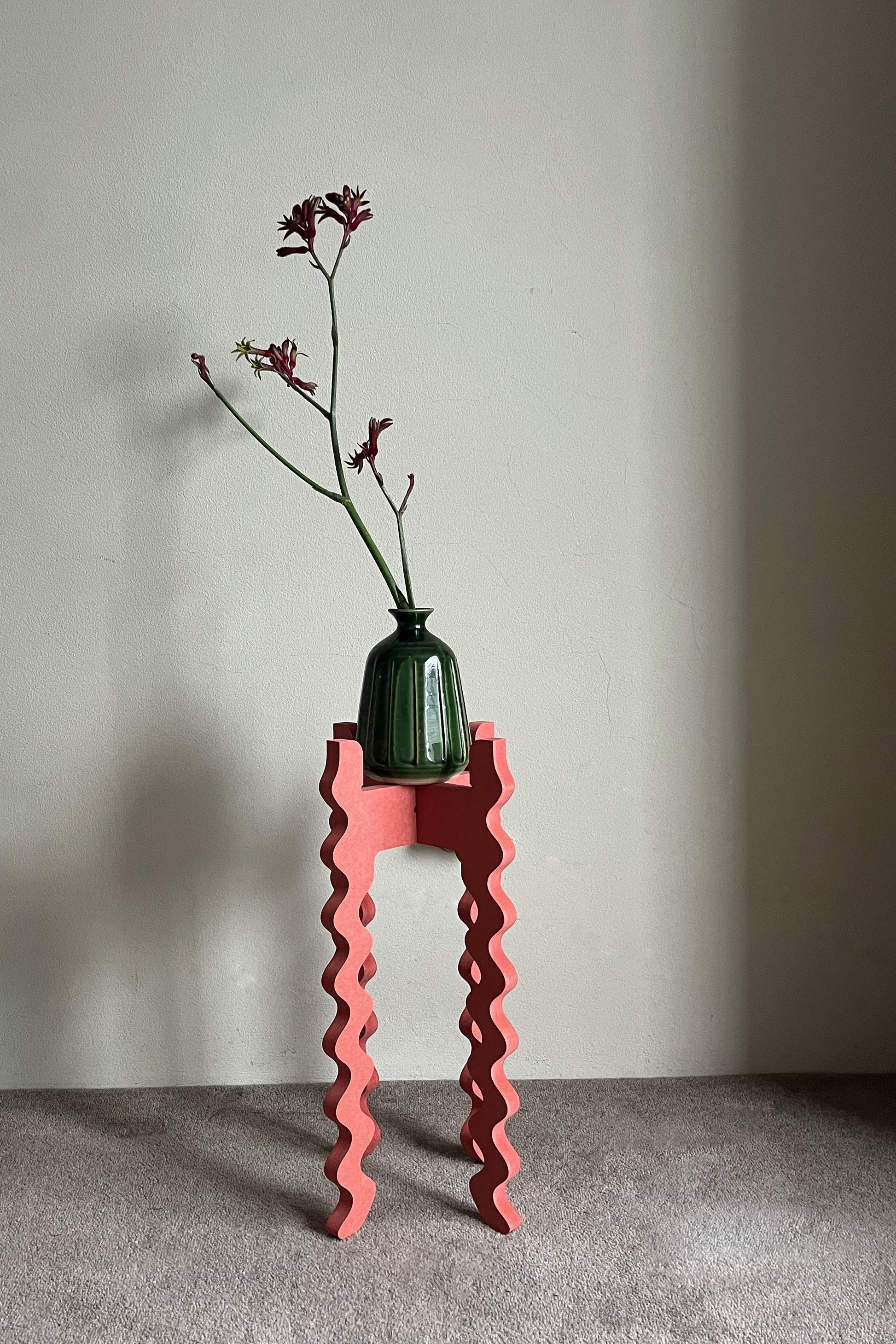 Green vase with a plant on a red wavy Xigglie Poppy Tallboy Wide Plant Stand against a plain wall.