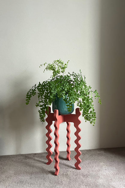 Green potted plant on a red wavy Xigglie Poppy Tallboy Wide Plant Stand against a plain wall.