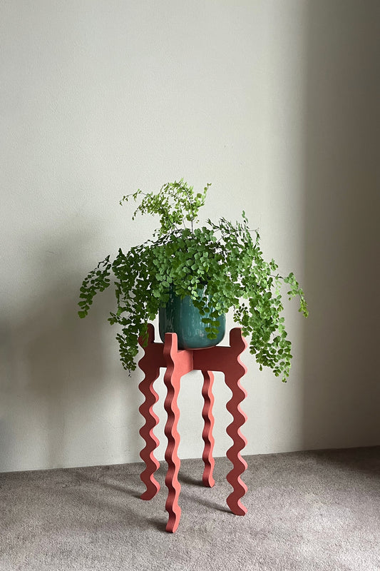 Green potted plant on a red wavy Xigglie Poppy Tallboy Wide Plant Stand against a plain wall.