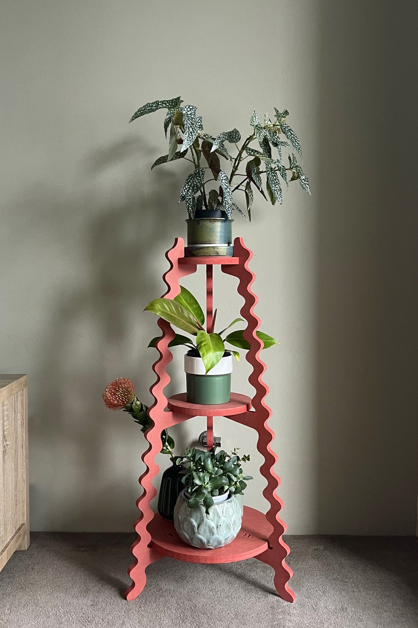 A red wavy Xigglie Poppy Threezy Plant Stand  supporting potted plants on three tiers against a neutral wall.
