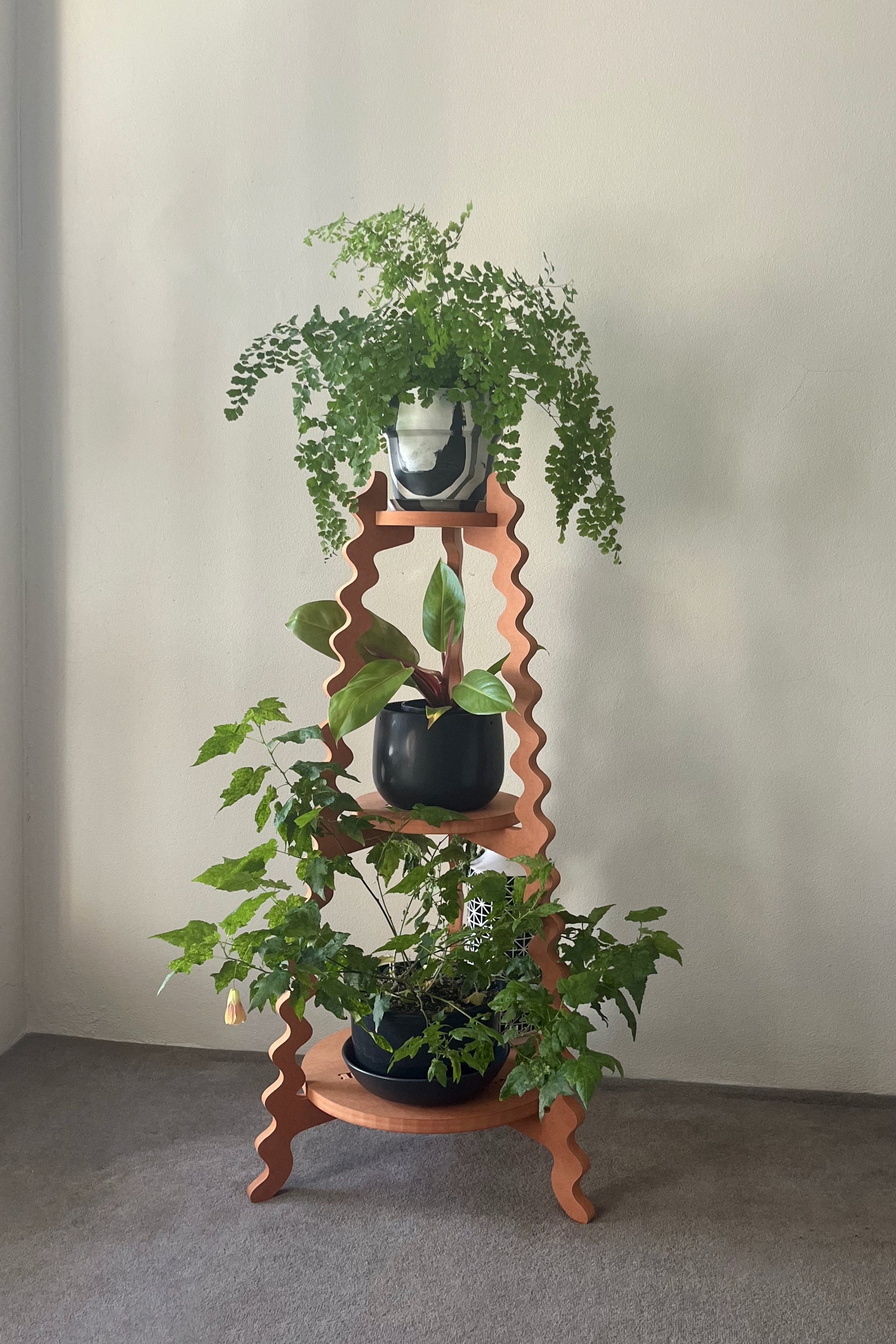 A orange wavy Xigglie Mango Threezy Plant Stand  supporting potted plants on three tiers against a neutral wall.