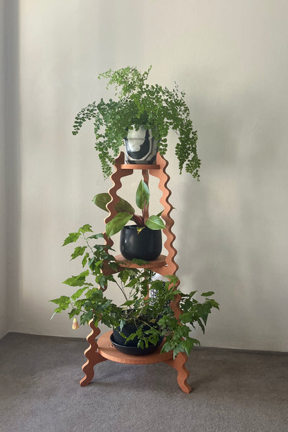A orange wavy Xigglie Mango Threezy Plant Stand  supporting potted plants on three tiers against a neutral wall.