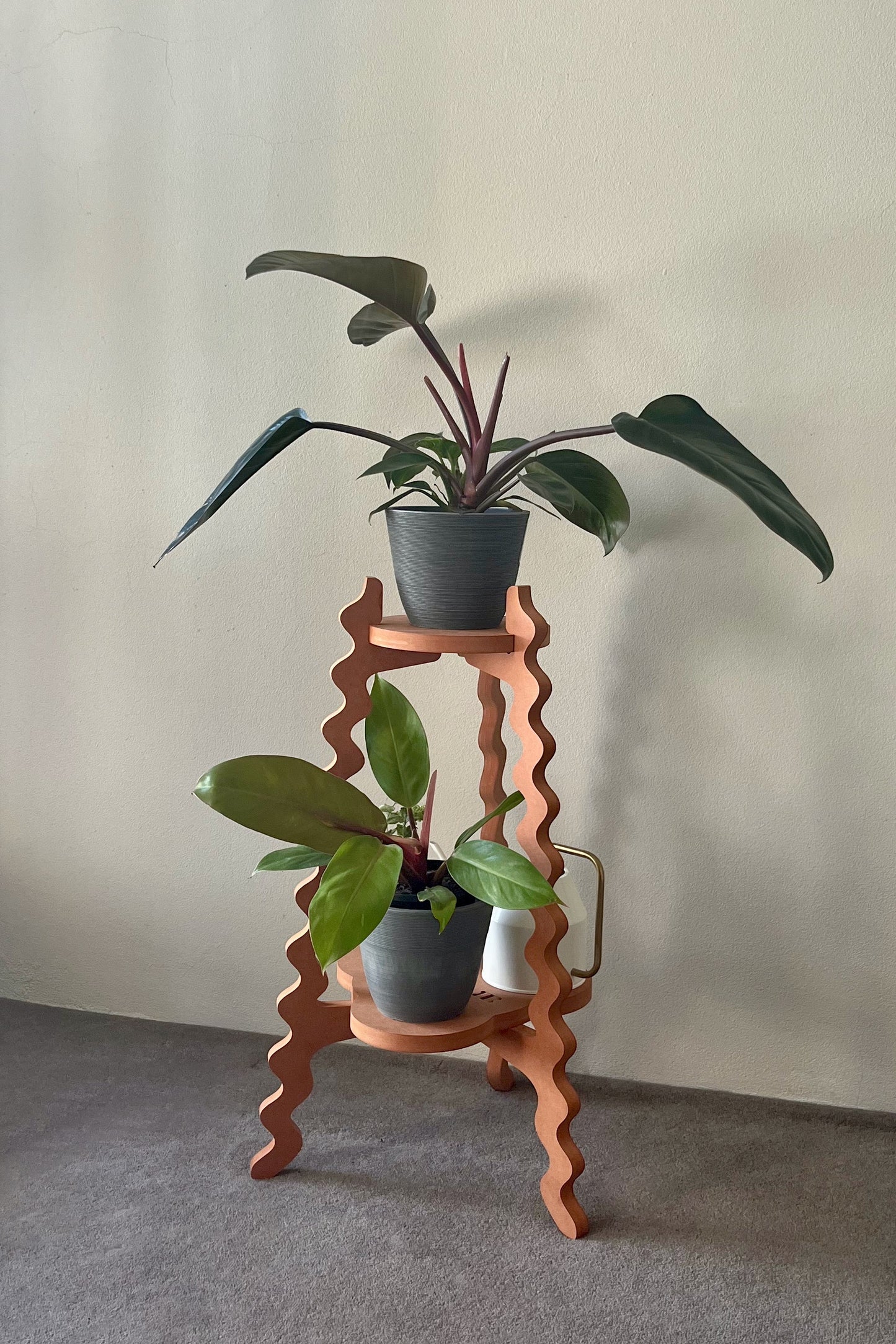Orange wavy Xigglie Mango Two-UP Plant Stand with a vase of flowers and potted plants against a plain wall.