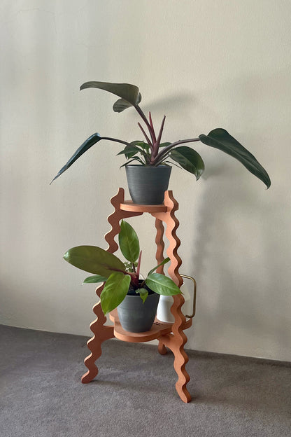 Orange wavy Xigglie Mango Two-UP Plant Stand with a vase of flowers and potted plants against a plain wall.