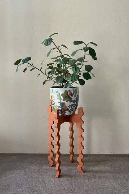 Green potted plant on an orange wavy Xigglie Mango Tallboy Wide Plant Stand against a plain wall.