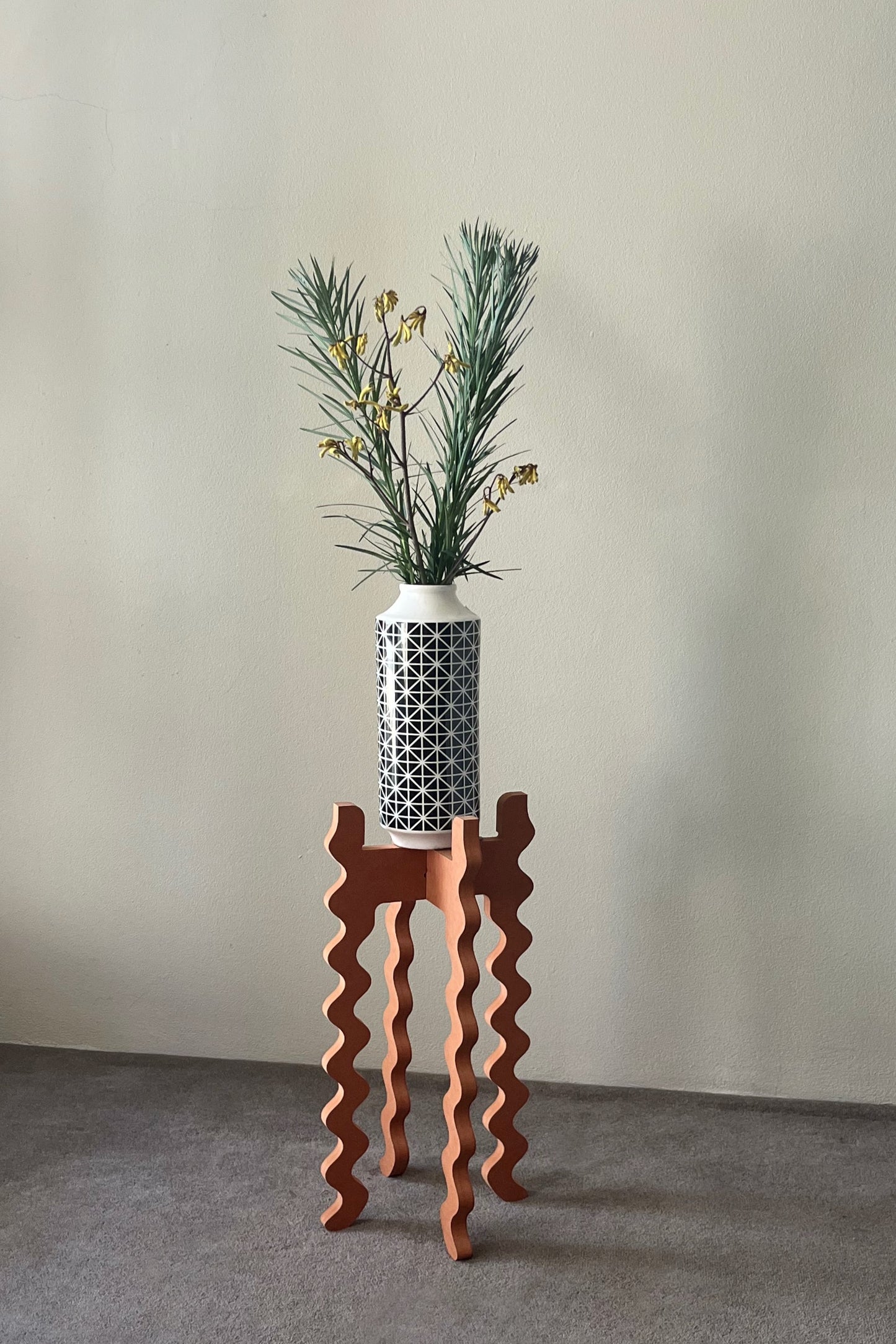 A black & white vase with flowers on an orange wavy Xigglie Mango Tallboy Wide Plant Stand against a plain wall.