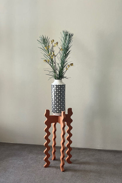 A black & white vase with flowers on an orange wavy Xigglie Mango Tallboy Wide Plant Stand against a plain wall.