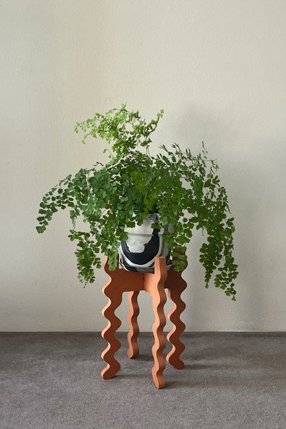 Potted plant on an orange wavy Xigglie Mango Middie Skinny Plant Stand against a plain wall