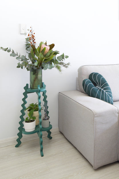 A Xigglie Sage Two-Up Plant Stand. Australian designed and made in green moisture-resistant MDF, supporting a vase of flowers and plants in pots, in a modern living room.