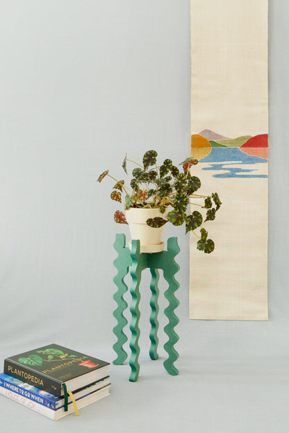 A Xigglie Sage Tallboy Skinny Plant Stand. Australian designed and made in green moisture-resistant MDF, supporting plant in ceramic pot, beside stack of books.