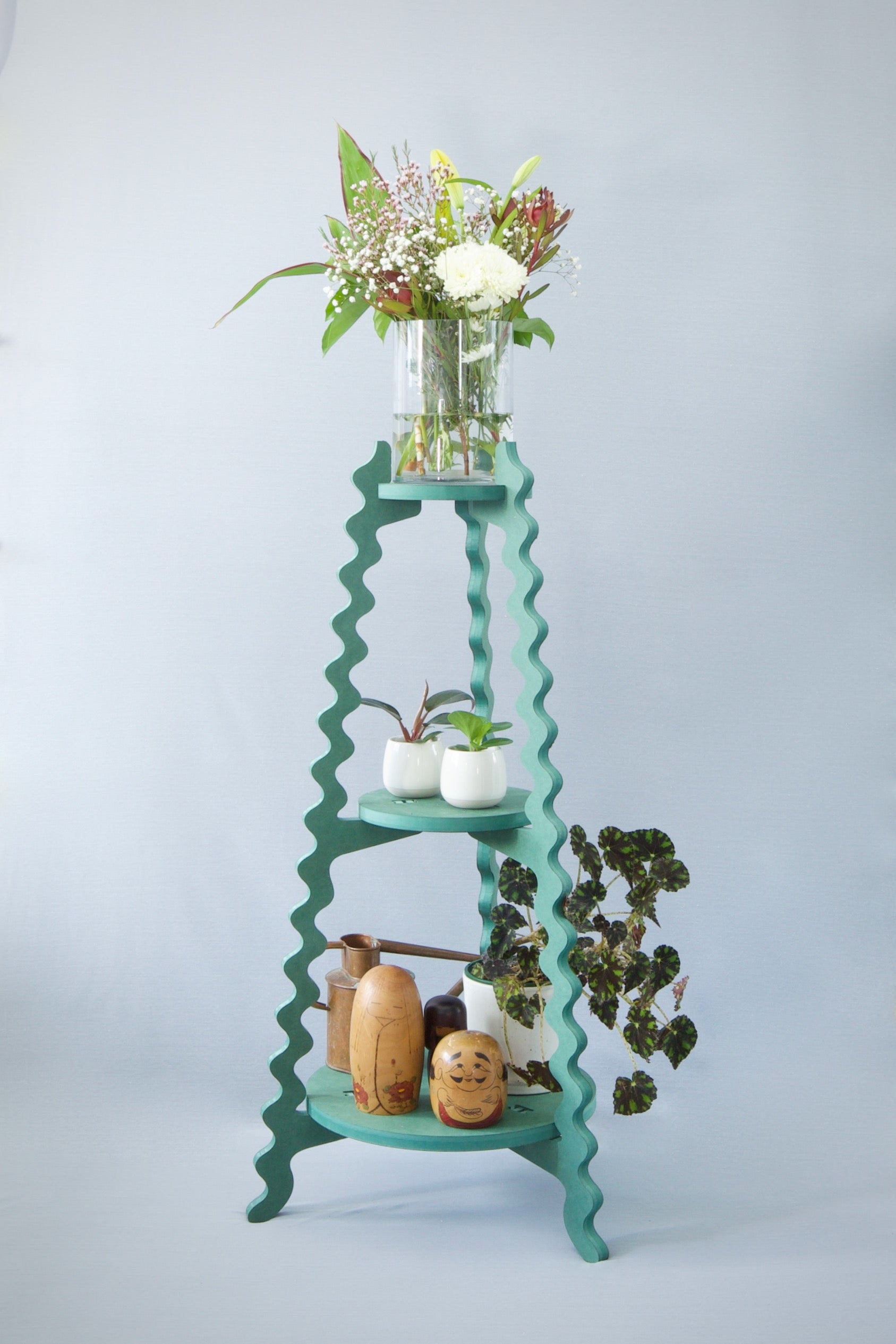 A Xigglie Sage Threezy Plant Stand. Australian designed and made in green moisture-resistant MDF, 3 tiers supporting flowers in vase and plants in pots.