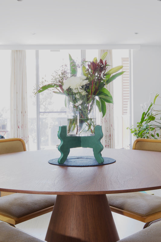 A Xigglie Sage Low-Rider Plant Stand. Australian designed and made in green moisture-resistant MDF, supporting flowers in vase at centre of round dining table.